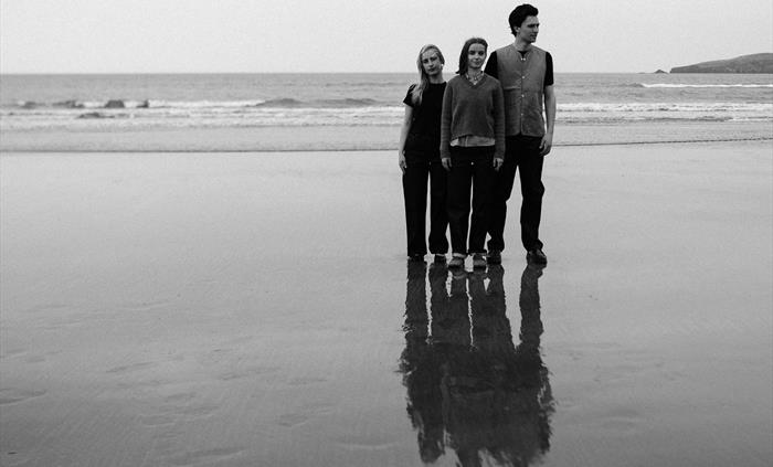 Three people stand on a wet beach facing slightly to the right, their reflections mirrored in the sand. The black-and-white photo captures a calm, coa